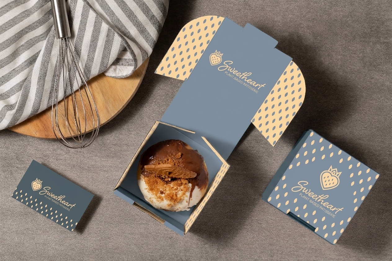 Branded blue-gray pastry packaging surrounds an open box containing a round iced pastry beside a whisk and striped napkin.